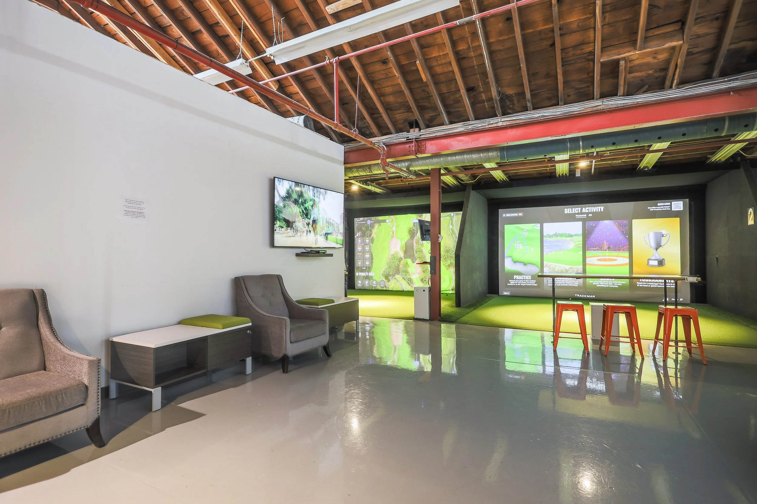 A wide hitting room with two projection screens, a high table with orange stools, and lounge seating along the left wall.