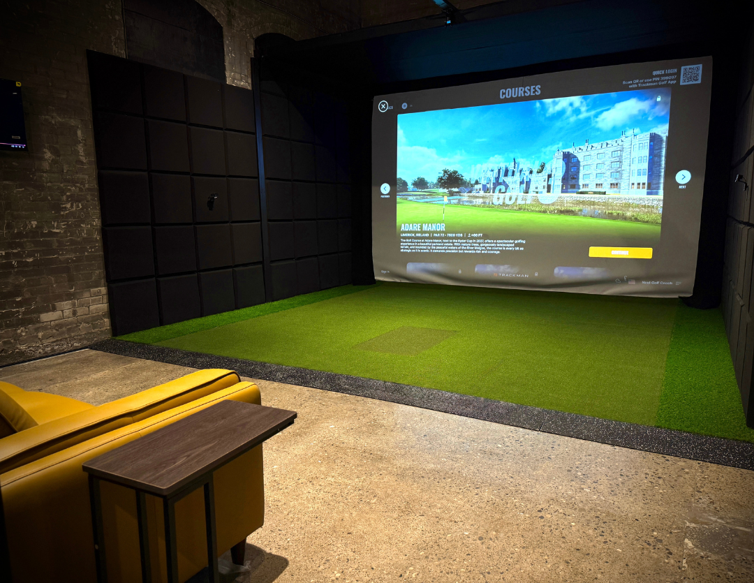 A private hitting room with dark acoustic wall panels, a course-selection menu on the screen and a green turf tee area.